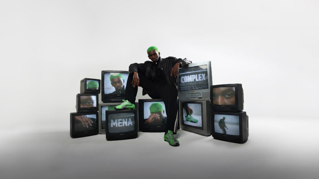 A male model with bright green hair wearing a black leather jacket and green sneakers sits on stacked vintage television sets, with screens displaying his face and the words “Complex” and “MENA” against a white studio background.