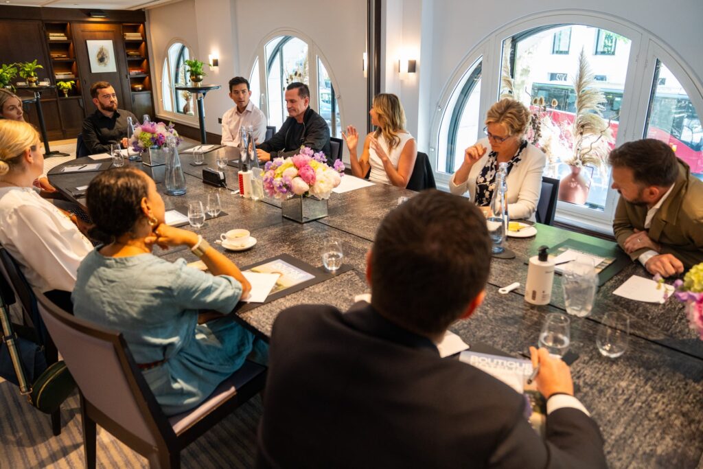 A group of hospitality professionals seated around a long dining table in a bright room, discussing ideas while notebooks, water glasses, and floral centerpieces sit on the table in front of them.