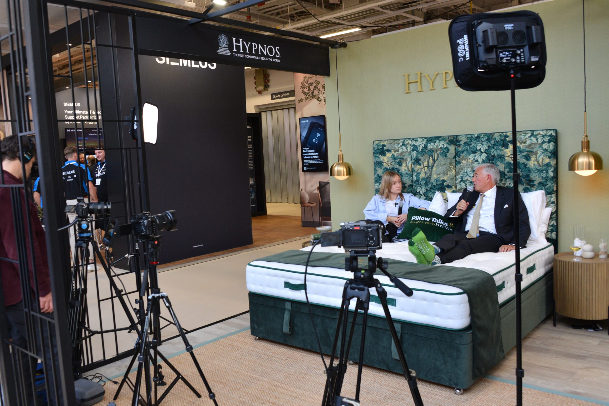 A professional video interview setup inside a hotel trade show booth, where a man in a suit and a woman sit on a large display mattress speaking to each other while multiple cameras, tripods, and studio lights record the conversation.