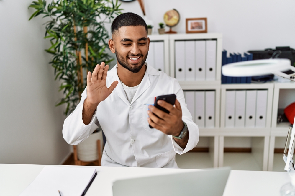 A man wearing a white shirt sitting at a desk, smiling and waving while holding a smartphone during a video call, with office folders and a plant visible behind him.