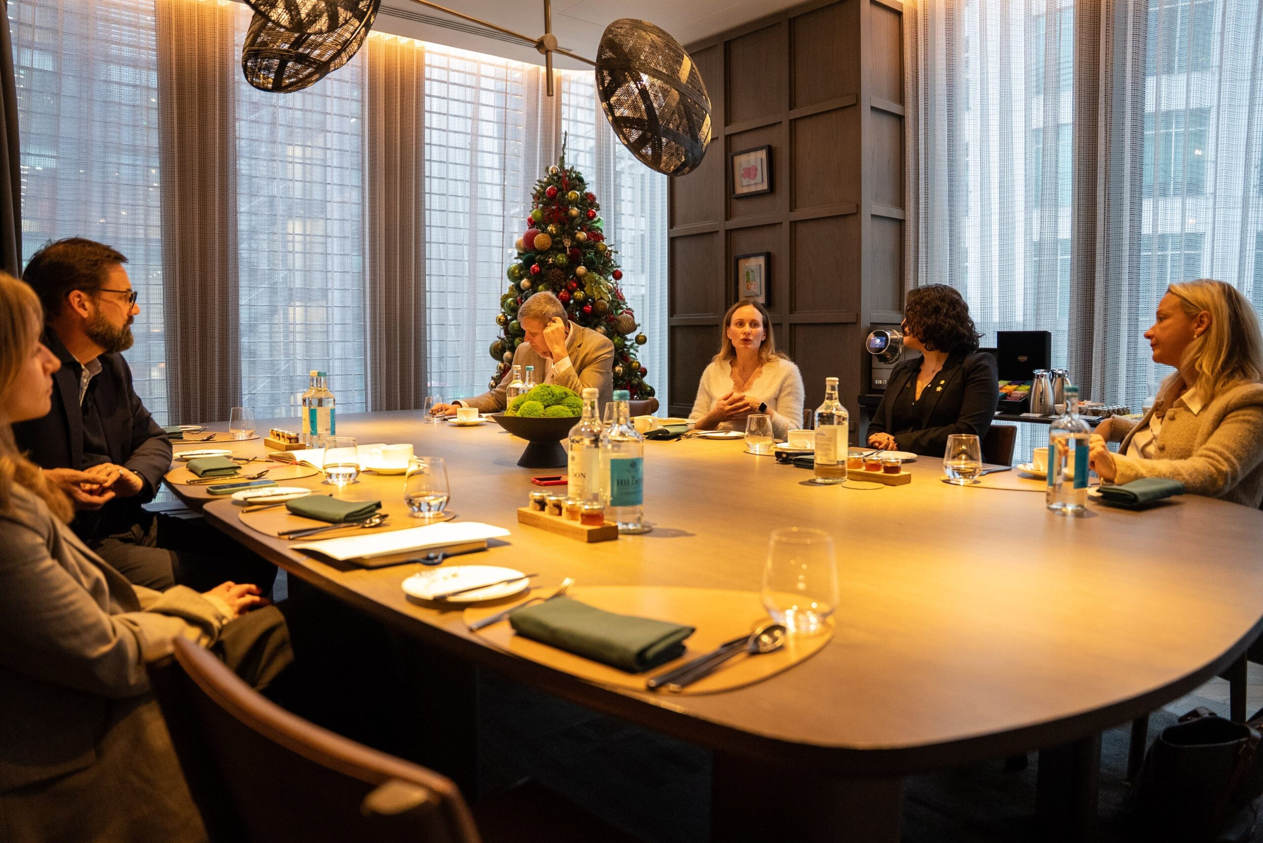 A group of hospitality professionals seated around a large oval wooden table in a modern meeting room, discussing ideas while notebooks, water bottles, wine glasses, and place settings are arranged in front of them, with floor-to-ceiling windows and a decorated Christmas tree in the background.
