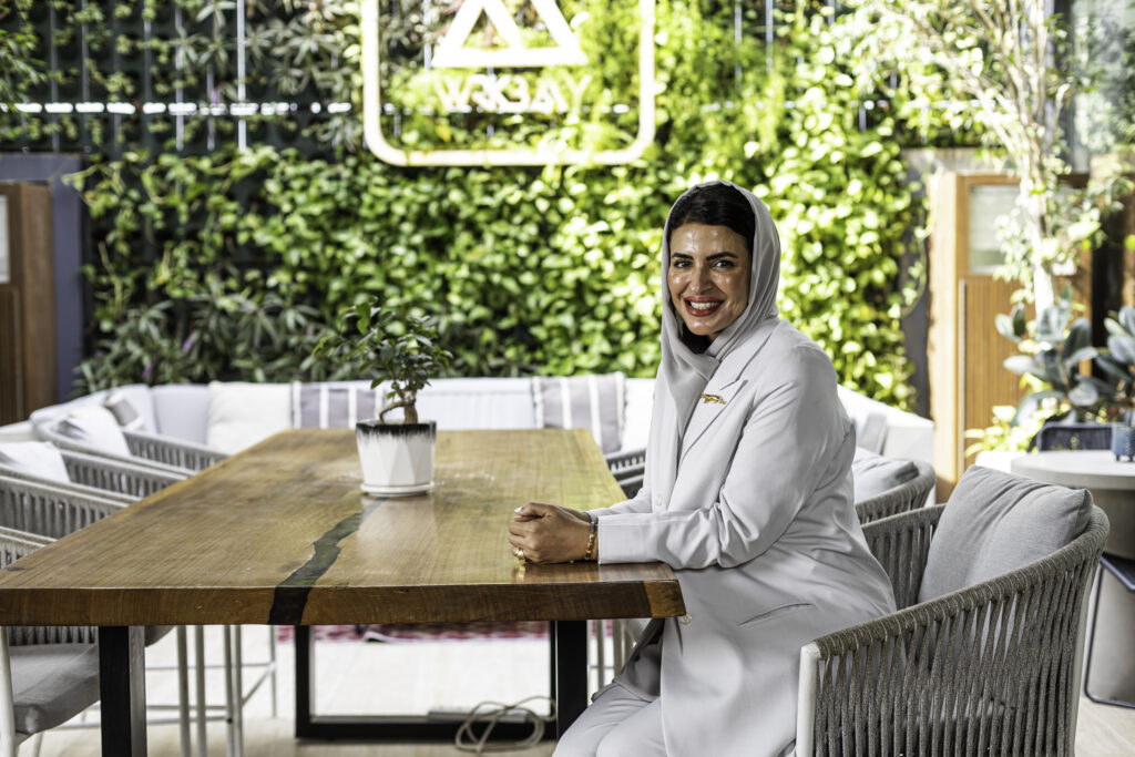 Noor Al Muhaideb seated at a wooden table in a bright outdoor setting with greenery behind her, smiling while engaging with the camera.