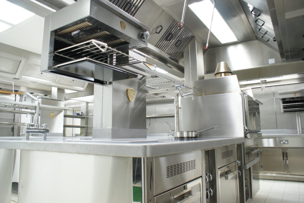 A modern stainless steel commercial kitchen with industrial cooking equipment, ventilation systems, shelving, and a metal frying pan resting on a cooktop.