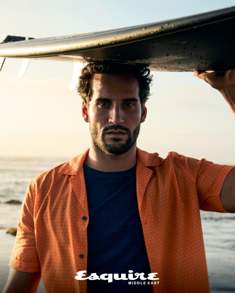 Male model holding a surfboard at the beach wearing a casual summer outfit for an Esquire Middle East fashion editorial.