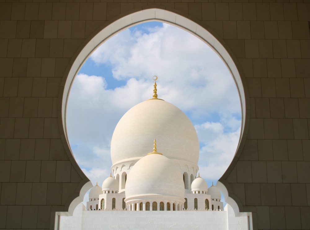 A large white mosque dome framed by an architectural archway, with smaller domes surrounding it and a blue sky visible in the background.