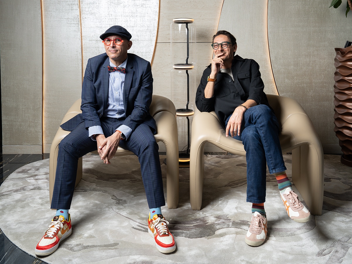Two well-dressed men seated side by side on chairs in a studio setting, wearing smart casual outfits and posing confidently for a magazine feature.
