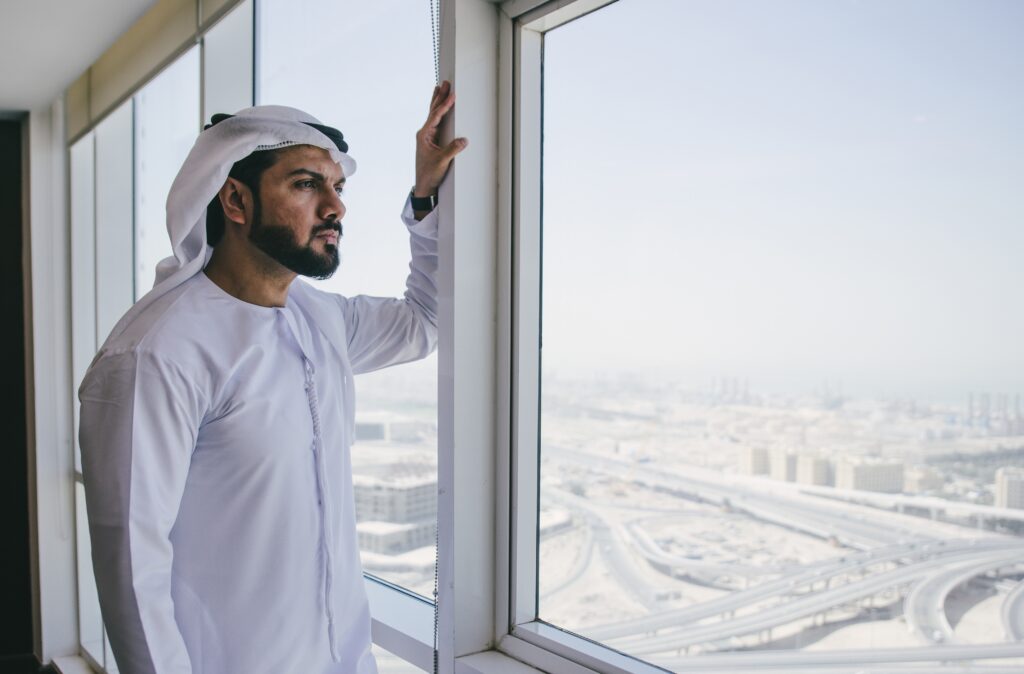 A Middle Eastern man wearing a white kandura and headscarf stands beside a large window in a high-rise building, looking out over a city skyline with a thoughtful expression.